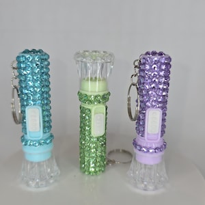 Może przedstawiać: Trzy błyszczące latarki w kolorze turkusowym, zielonym i fioletowym. Każda latarka ma przezroczystą, fasetowaną podstawę i srebrny brelok do kluczy. Turkusowe i fioletowe latarki mają pasujące kolorystycznie klejnoty.