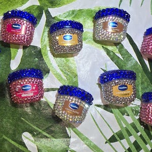 May include: Eight small Vaseline lip therapy jars, each featuring a blue rhinestone-encrusted lid and a body covered in either pink or clear rhinestones. The labels display various product names including Rosy Lips, Creme Brulee, and Mistletoe Mint.