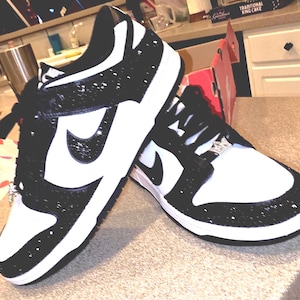 May include: Pair of black and white sneakers with a sparkling glitter design. The shoes feature a white toe box, a black Nike swoosh, and black laces. The sneakers are decorated with rhinestones.