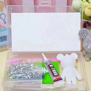 May include: A clear plastic storage box with a hinged lid, filled with craft supplies. Contents include rhinestones, glue, a white bear figurine, and other small items. The glue tube has the text "B-7000" on it.