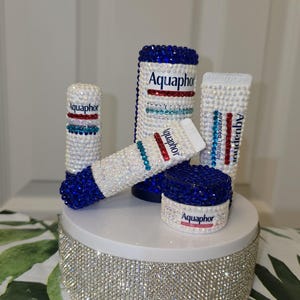 Puede incluir: Una colección de productos Aquaphor, incluyendo tubos y tarros, adornados con brillantes pedrería. Los productos están dispuestos sobre una plataforma blanca cubierta de pedrería, creando una exhibición glamurosa. El texto "Aquaphor" es visible en cada artículo.