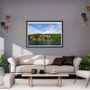 May include: A framed photograph of a rocky coastline with a winding road leading up the side of the cliff. The photo is hanging on a gray wall above a beige sofa with brown throw pillows.