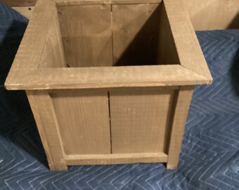 Könnte beinhalten: Ein quadratischer Holz-Pflanzkasten mit einer natürlichen braunen Oberfläche. Der Pflanzkasten hat ein schlichtes Design mit einem breiten Rand und einer glatten Oberfläche.