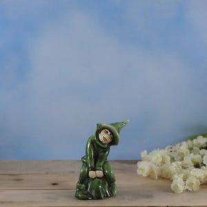 Op de afbeelding: Een kleine, groene keramische figuur van een persoon in een capuchon en hoed. Het beeldje heeft een lachend gezicht en zit. De achtergrond is een lichtblauwe lucht en een houten oppervlak.