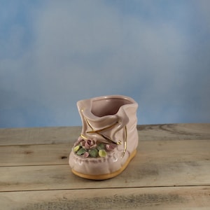 Könnte beinhalten: Ein rosafarbener Keramik-Babyschuh-Pflanztopf mit goldenen Akzenten. Der Schuh ist mit einem Cluster aus rosa und grünen floralen Verzierungen verziert. Er hat eine hellbraune Sohle und steht auf einer Holzoberfläche.