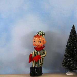Könnte beinhalten: Eine Elfenfigur im Vintage-Stil, die ein grün-weiß gestreiftes Outfit mit passender Zipfelmütze, rotem Kragen, roten Fäustlingen und schwarzen Stiefeln trägt. Der Elf hat ein bemaltes Gesicht mit rosigen Wangen und steht aufrecht da.