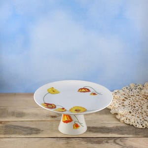 May include: A white ceramic cake stand featuring a painted floral pattern with yellow and orange poppy-like flowers and thin green stems. The stand has a circular flat top and a flared pedestal base, showcasing a smooth, glossy finish throughout.