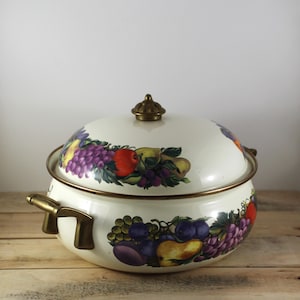 May include: Cream-colored enamelware pot with lid, adorned with a colorful fruit design featuring grapes, apples, and pears. The pot has brass-toned handles and a matching knob on the lid. The pot sits on a wooden surface.