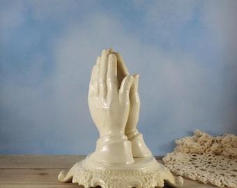 May include: Ivory ceramic sculpture of praying hands. The hands are clasped together, with detailed fingers and a smooth finish. The base is ornate with a decorative, scrolled design. The sculpture is set against a light blue background.
