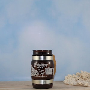May include: A brown glass mug with a silver band and a wooden handle. The mug has the words "DOGPATCH U.S.A." in white lettering, along with a graphic of cartoon characters. The mug also has the text "God is better than Evil because it's nicer!"