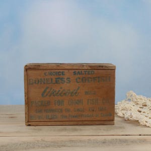 Könnte beinhalten: Eine verwitterte Holzkiste mit der Aufschrift "CHOICE SALTED BONELESS CODFISH Unicoid BRAND PACKED FOR UNION FISH CO." in verblichener grüner Schrift. Die rechteckige Kiste steht auf einer Holzoberfläche.