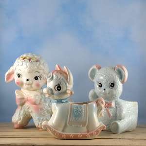 Può includere: Tre statuine in ceramica: un agnello, una fioriera a forma di cavallo a dondolo e un orsacchiotto. L'agnello e l'orsacchiotto sono bianchi e azzurri con accenti rosa. Il cavallo a dondolo è azzurro e bianco con dettagli rosa.