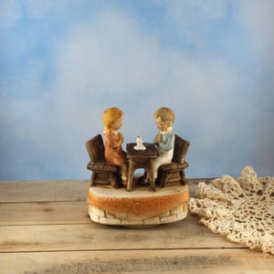 May include: A ceramic figurine of two children seated at a small table with a lit candle. The children are dressed in vintage clothing and seated on wooden chairs. The figurine has a light brown base with a decorative border.