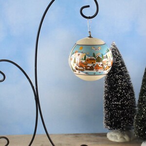 Peut inclure: Une boule de Noël vintage avec une scène de village d'hiver, suspendue à un support en métal noir. L'ornement présente un fond bleu clair avec un village peint, des maisons et des arbres. L'année "1979" est visible sur l'ornement.