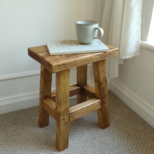May include: A small, wooden stool with a square top and four legs. A book and a grey mug rest on the stool. The stool is made of light brown wood and is placed on a patterned carpet. The stool's height is approximately 40 cm.