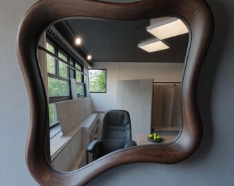 May include: A large, uniquely shaped mirror with a dark brown, wavy wooden frame. The mirror reflects a room with a black leather chair, windows, and a table with a bowl of green fruit. The mirror is mounted on a gray wall.