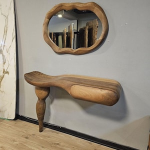 Könnte beinhalten: Ein Wandspiegel aus Holz mit einer einzigartigen, organischen Form. Der Spiegel hat ein einzelnes, geschwungenes Bein und ein Regal. Ein Holzspiegel mit einem unregelmäßigen, wellenförmigen Rahmen hängt über dem Spiegel.