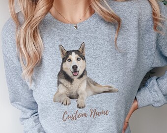 Op de afbeelding: Een grijs sweatshirt met een bedrukte Siberische Husky-hond die ligt met zijn poten voor zich. De tekst "Custom Name" is onder de hond gedrukt.