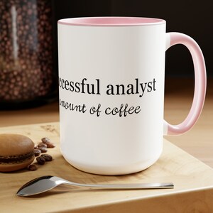 May include: White ceramic coffee mug with a pink rim and handle. The mug has black text that reads "A successful analyst needs a substantial amount of coffee".