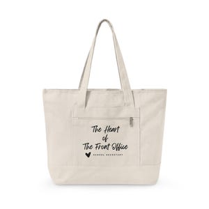 Op de afbeelding: Een crèmekleurige canvas tote bag met twee schouderbanden en een voorvak met ritssluiting. Op het voorvak staat de zwarte tekst "The Heart of The Front Office" gedrukt, gevolgd door een klein harticoontje en de woorden "SCHOOL SECRETARY".