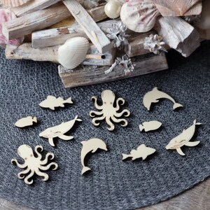Könnte beinhalten: Holz-Ausschnitte verschiedener Meeresbewohner, darunter ein Oktopus, ein Wal, ein Delfin und Fische, auf einer grauen, strukturierten Oberfläche.