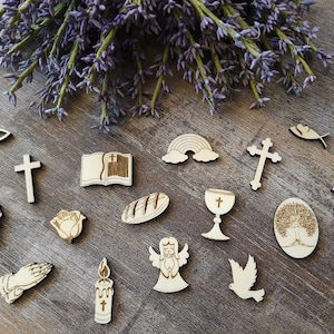 Könnte beinhalten: Holzschnitzereien von religiösen Symbolen, darunter ein Kreuz, ein Kelch, ein Fisch, eine Taube, ein Buch, eine Kerze, eine Weinrebe, eine Rose, ein Brotlaib, ein Regenbogen, ein Baum und das Chi-Rho-Symbol. Die Schnitzereien sind auf einer Holzoberfläche mit Lavendelzweigen angeordnet.
