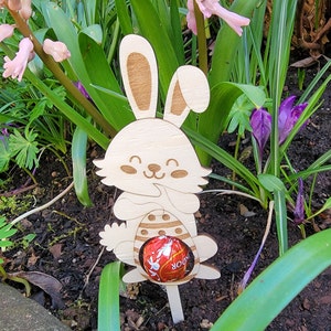 Peut inclure: Découpe de lapin de Pâques en bois tenant un œuf en chocolat. Le lapin est peint en blanc avec des oreilles brunes et un œuf brun avec des rayures blanches. Le lapin sourit et a un visage mignon.