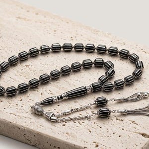 Può includere: Una corona di preghiera con perline sfaccettate a forma di botte a righe bianche e nere. Include una perla centrale lunga e affusolata a righe, dettagli in metallo color argento e due catenelle color argento che terminano con delle nappe.