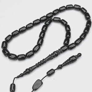 Può includere: Una corona di grani da preghiera composta da perline cilindriche nere, lisce e lucide, disposte in un cerchio continuo. Il filo include un elemento centrale decorativo intagliato nero e una sezione a nappa con perline nere aggiuntive.