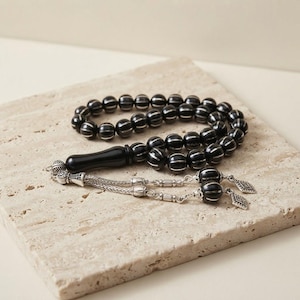 Può includere: Una corona di grani da preghiera con perle sferiche nere e intarsi verticali color argento. Include una perla focale nera liscia e allungata e un componente a nappa in metallo color argento con due piccoli ciondoli a forma di rombo.