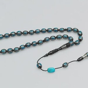 May include: A string of prayer beads featuring black oval beads decorated with blue and white eye motifs. The strand includes a long black cylindrical piece with silver rings, two smaller black beads, and a single rectangular turquoise stone.