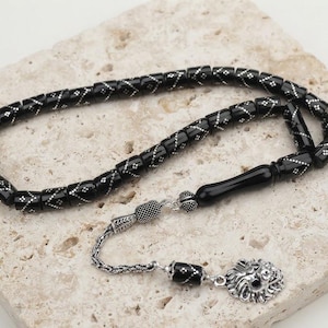 Può includere: Una corona di grani da preghiera composta da perline cilindriche nere con intricati motivi intarsiati color argento. Il filo include una perla centrale nera liscia, distanziatori in metallo argentato e un ciondolo a forma di testa di leone.