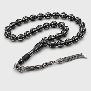 May include: A prayer bead strand featuring glossy black spherical beads with vertical silver-toned stripes. The strand includes a tapered black handle piece and a decorative silver-toned metal tassel with intricate braided chain detailing.