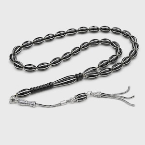Peut inclure: Un chapelet composé de perles ovales noires avec de fines rayures verticales blanches. Le collier comprend une perle centrale allongée noire et blanche, un connecteur en métal argenté et une chaîne à pampilles décorative argentée.