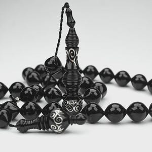 May include: A set of black prayer beads featuring faceted, glossy beads strung on a black cord. The central pendant and the terminal piece are decorated with intricate silver-colored filigree patterns, providing a contrast to the dark beads.