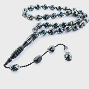 Può includere: Una corona di grani da preghiera con perle nere di forma ovale decorate con motivi a vortice bianchi e accenti blu. Il filo include un lungo elemento centrale nero scanalato e un cordino nero che termina con due perle più piccole coordinate.