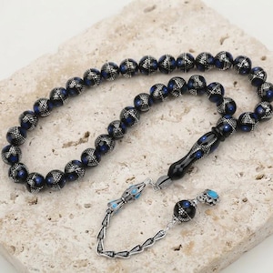 Può includere: Una corona di grani da preghiera con perle sferiche blu scuro decorate da intricati motivi in filigrana color argento. Il filo include una perla nera allungata più grande e una nappa decorativa in metallo color argento con accenti blu.