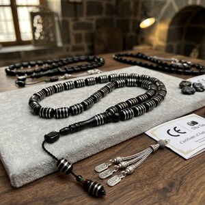 May include: A prayer bead strand featuring cylindrical black beads with silver-coloured metallic bands. The strand includes a black cord, a decorative silver-coloured tassel with three engraved metal charms, and a certificate of authenticity card.