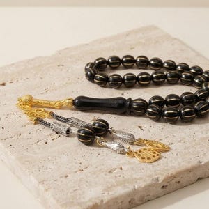 Op de afbeelding: Een gebedsketting met zwarte, bolvormige kralen voorzien van dunne, goudkleurige verticale strepen. De streng bevat een gladde, langwerpige zwarte kraal, een goudkleurige metalen verbinding en decoratieve zilver- en goudkleurige metalen bedels.