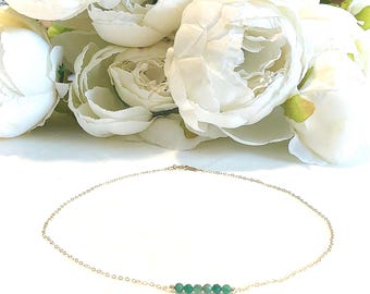 May include: A delicate gold necklace with a strand of small, green beads. The necklace is set against a backdrop of soft, white flowers, creating a simple and elegant aesthetic. The necklace is a choker style.