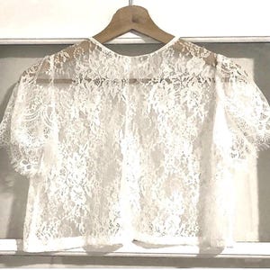 Puede incluir: Una blusa de encaje blanco con mangas cortas, colgada de una percha de madera. El delicado patrón floral de encaje cubre toda la blusa, creando un efecto semitransparente. La blusa se muestra sobre un fondo blanco.