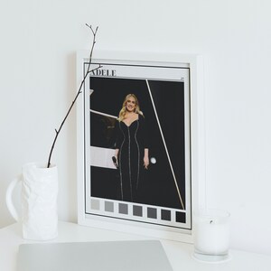 Puede incluir: Una foto en blanco y negro enmarcada de Adele con un vestido negro, con la palabra "ADELE" impresa en blanco en la parte superior de la foto. La foto está rodeada de un marco blanco y una fila de cuadrados grises en la parte inferior.