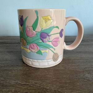 May include: A pink ceramic mug with a floral design featuring purple, pink, and yellow tulips. The mug has a light pink handle and a decorative base with a brick pattern. The background is a light blue wall and a wooden surface.