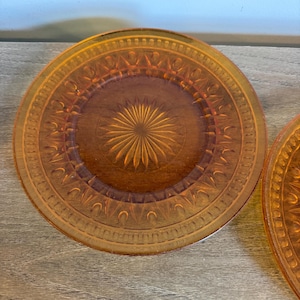 May include: A round amber-colored glass plate featuring a central sunburst pattern surrounded by a ring of embossed arches and a textured outer rim. The translucent material catches the light, highlighting the intricate geometric pressed glass design.