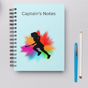 May include: A light blue spiral-bound notebook with the text "Captain's Notes." The cover features a black silhouette of a field hockey player against a colourful splash of orange, pink, and blue. A white pencil and blue pen are next to the notebook.
