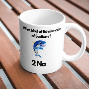 May include: White ceramic mug with a blue fish illustration and the text "What kind of fish is made of Sodium? 2Na".
