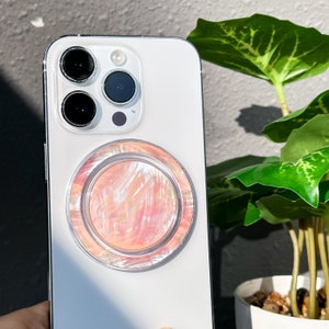 May include: A white iPhone with a pink and white iridescent phone ring attached to the back.