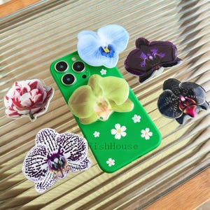 May include: A green phone case adorned with white flower accents and several orchid-shaped phone grips in various colors, including blue, purple, black, and a red and white flower. The text "FishHouse" is visible on the case.