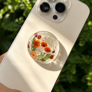 May include: A circular phone grip attached to a light-coloured smartphone. The grip is made of clear resin containing pressed orange flowers, green foliage, and small gold flakes. The text FlyFishHouse is printed on the phone surface.