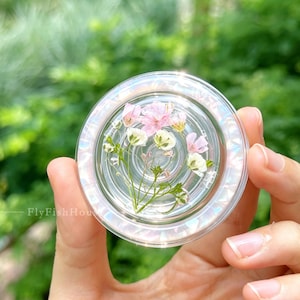 May include: A circular phone grip featuring a clear, iridescent resin casing with embedded pressed flowers. The floral arrangement includes small pink and white blossoms with green stems and leaves. The text FlyFishHouse is printed on the left.